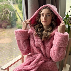 Può includere: Un morbido accappatoio con cappuccio rosa con tessuto spesso e strutturato. L'accappatoio presenta un collo a scialle, ampi polsini e una cintura abbinata. La persona è seduta su una sedia di legno vicino a una finestra.