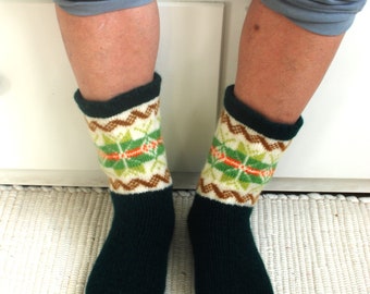May include: A pair of dark green socks with a white, brown and green geometric pattern. The socks are being worn by a person sitting on a white bench.