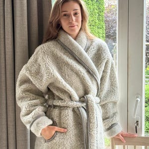 May include: A light gray, plush robe with a shawl collar and a matching belt. The robe has gray trim along the edges and cuffs. The model is wearing the robe and standing near a window.