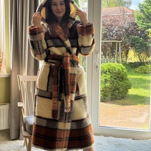 May include: A hooded, plaid robe in shades of cream, brown, and orange. The robe has a matching belt and is made of a soft, textured material. The person is wearing black boots. The robe is designed for comfort and warmth.