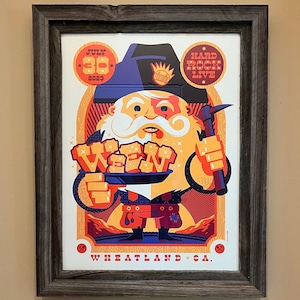 May include: A framed poster with a cartoon illustration of a bearded man wearing a blue hat and holding a pickaxe and a plate with the word "WEEN" on it. The poster is red, orange, yellow, and blue. The text on the poster reads "JULY 30 2023 HARD ROCK LIVE WEEN WHEATLAND * CA."