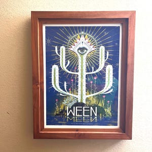 May include: Framed art print featuring a stylized cactus with an eye at the top, set against a dark blue background with radiating lines. The print includes the band name "WEEN" in white, with a brown wooden frame.