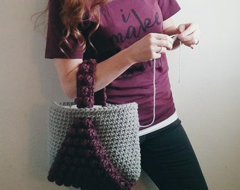 May include: A woman is crocheting a grey and purple tote bag with a braided handle. The bag has a textured design and a large purple triangle on the front. The woman is wearing a maroon t-shirt with the text "i make" on it.