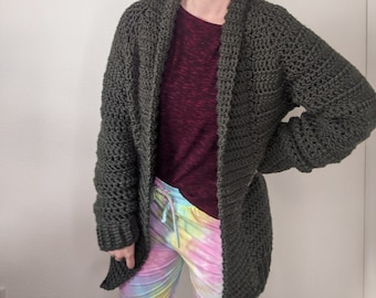 May include: A woman wearing a dark green crocheted cardigan with a long, open front and long sleeves. She is wearing a maroon t-shirt and tie-dye trousers.