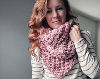 May include: A hand-knitted cowl in a soft, dusty rose colour. The cowl is made of thick yarn, creating a textured, warm accessory. The cowl is worn over a striped long-sleeve top and blue jeans.