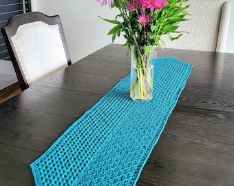 May include: A teal crochet table runner with a geometric openwork pattern. A clear glass vase holds a bouquet of pink, purple, and green flowers. The runner is on a dark wood table, with two upholstered chairs in the background.