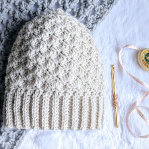 Könnte beinhalten: Eine gehäkelte Mütze in hellbeige mit einem strukturierten Stichmuster. Die Mütze liegt auf einer weißen Oberfläche mit einer Häkelnadel und einem Maßband.
