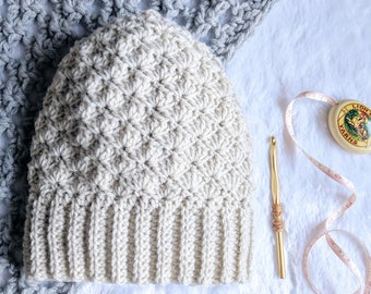 May include: A crocheted beanie hat in a light beige colour with a textured stitch pattern. The hat is lying on a white surface with a crochet hook and a measuring tape.