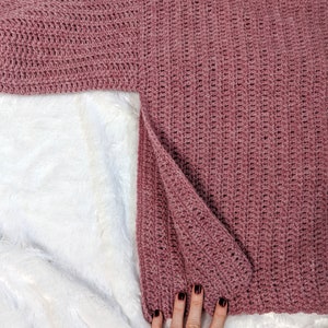 May include: A pink crocheted sweater with a long sleeve. The sweater is laid flat on a white fuzzy surface.