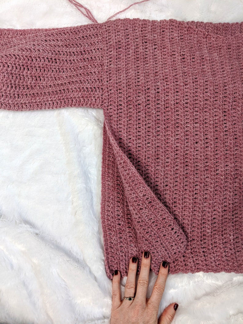 May include: A pink crocheted sweater with a long sleeve. The sweater is laid flat on a white fuzzy surface.