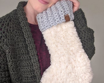 May include: A Christmas stocking with a grey knitted cuff and a cream-coloured, fluffy body. A small brown leather tag is attached to the cuff. The stocking is held by a person wearing a green knitted cardigan.