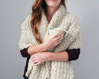 May include: A cream-coloured, chunky knit shawl. The shawl is draped over the shoulders. The texture of the knit is visible. The person is holding a light green mug. The background is a solid grey.