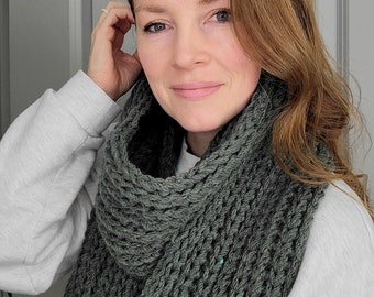 May include: A woman wearing a grey sweater and a dark green knitted infinity scarf. The scarf has a textured, open-weave design.