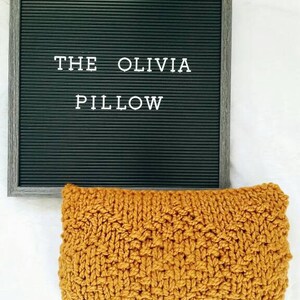 May include: A mustard yellow knitted pillow with the text "THE OLIVIA PILLOW" on a black letter board behind it.