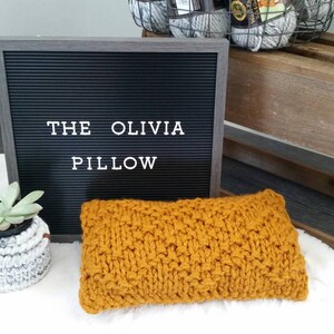 May include: A yellow knitted pillow with the text "THE OLIVIA PILLOW" on a black letter board.