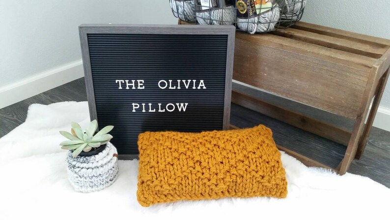 May include: A yellow knitted pillow with the text "THE OLIVIA PILLOW" on a black letter board.