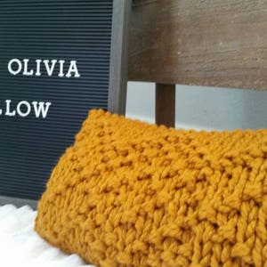 May include: A mustard yellow knitted pillow with a chunky knit texture. The pillow is sitting on a white surface with a black letter board behind it that says "THE OLIVIA PILLOW".