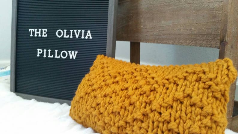 May include: A mustard yellow knitted pillow with a chunky knit texture. The pillow is sitting on a white surface with a black letter board behind it that says "THE OLIVIA PILLOW".
