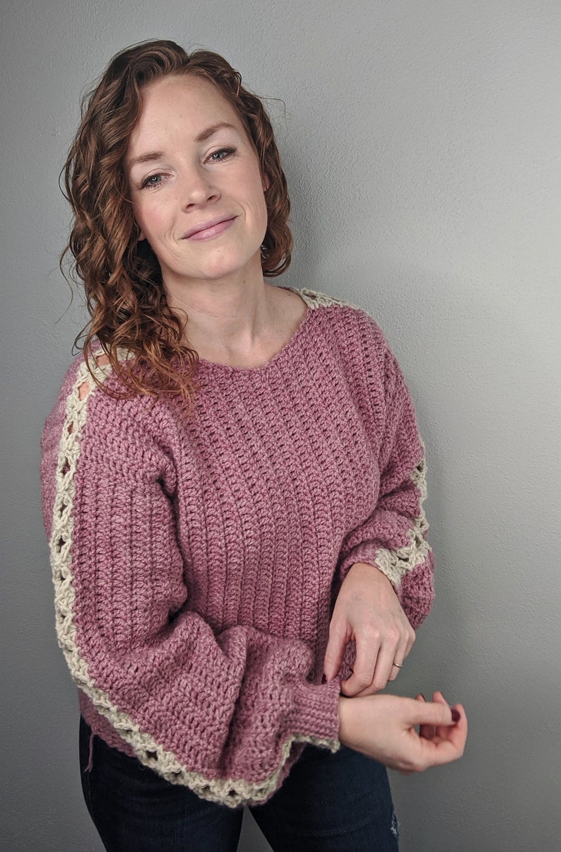 May include: A pink crocheted sweater with white crocheted trim on the sleeves. The sweater has a relaxed fit and a slightly cropped length.
