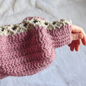May include: A pink crocheted sweater with a white crocheted trim around the cuffs. The sweater is made with a textured stitch.