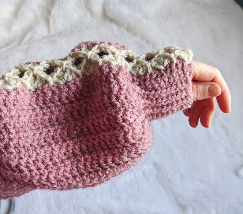 May include: A pink crocheted sweater with a white crocheted trim around the cuffs. The sweater is made with a textured stitch.