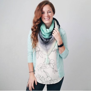 Puede incluir: Una mujer lleva una chaqueta de punto azul claro sobre una blusa blanca con estampado floral. Lleva una bufanda triangular de ganchillo de color turquesa, gris y blanco con flecos.