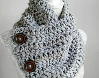 May include: A grey crocheted cowl with a button fastening. The cowl is made with a chunky yarn and has a simple, modern design.
