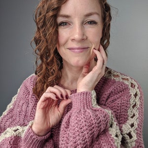 May include: A pink crocheted sweater with a white crocheted trim on the sleeves. The sweater has a relaxed fit and a slightly oversized look.