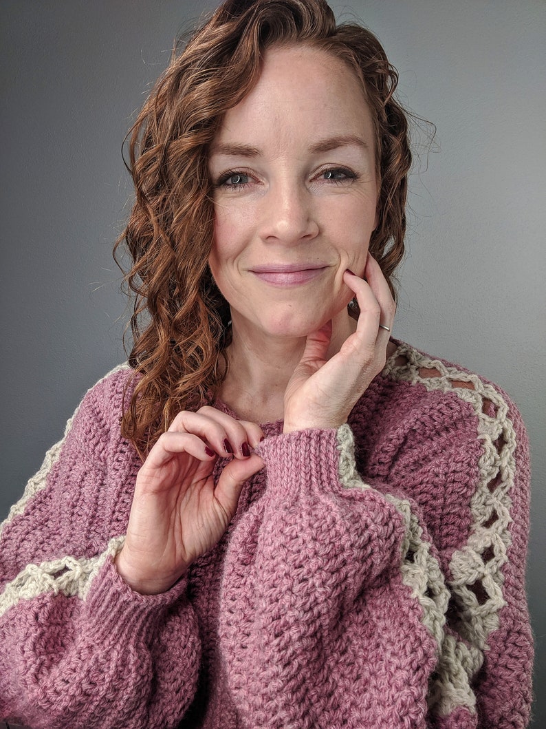 May include: A pink crocheted sweater with a white crocheted trim on the sleeves. The sweater has a relaxed fit and a slightly oversized look.