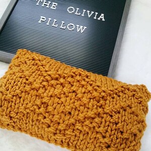 May include: A mustard yellow knitted pillow with a cable knit pattern. The pillow is sitting on a white surface with a black letter board that says "The Olivia Pillow".
