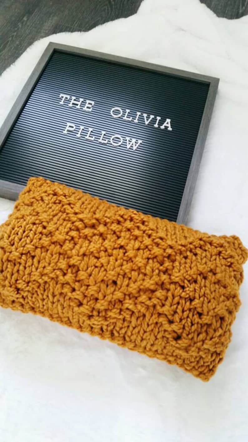 May include: A mustard yellow knitted pillow with a cable knit pattern. The pillow is sitting on a white surface with a black letter board that says "The Olivia Pillow".