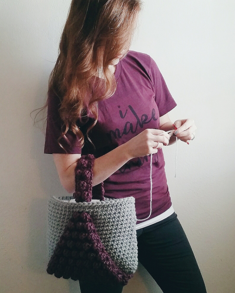 May include: A woman is crocheting a gray and purple tote bag with a braided handle. The bag has a textured design and a large purple triangle on the front. The woman is wearing a maroon t-shirt with the text "i make" on it.
