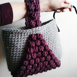 May include: A gray crocheted tote bag with a burgundy crocheted handle and a burgundy crocheted triangle design on the front.