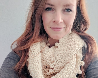 May include: A woman with long, wavy red hair is wearing a cream crocheted infinity scarf with a bobble stitch detail. The scarf is worn over a white blouse with a black floral pattern and a grey cardigan.