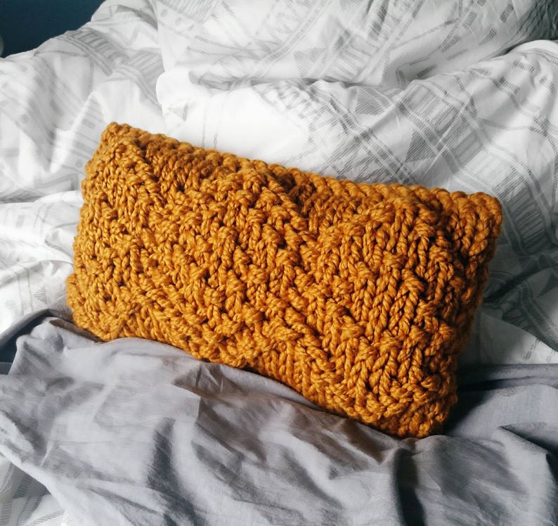 May include: A mustard yellow knitted pillow with a textured woven pattern. The pillow is lying on a gray bedspread.