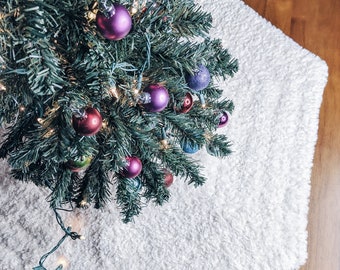May include: A white fluffy tree skirt with a hexagonal shape, placed on a wooden floor. A Christmas tree with green needles and colourful ornaments is positioned on top of the skirt.