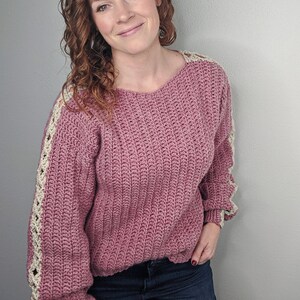 May include: A pink crocheted sweater with white crocheted trim on the sleeves. The sweater has a relaxed fit and a round neckline.