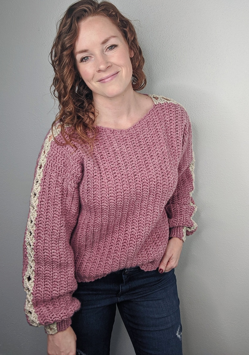 May include: A pink crocheted sweater with white crocheted trim on the sleeves. The sweater has a relaxed fit and a round neckline.