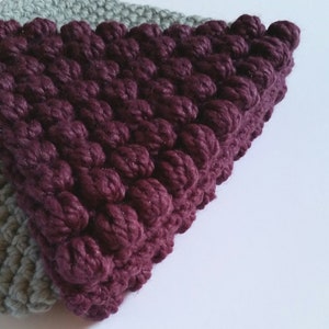 May include: Close-up of a knitted fabric with a textured, raised pattern. The fabric is a combination of two colors: a light gray and a deep burgundy. The burgundy portion of the fabric is in the shape of a triangle.