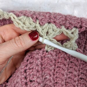 May include: Close-up of a person crocheting a pink and beige blanket. The person is holding a white crochet hook and is working on the beige border of the blanket.