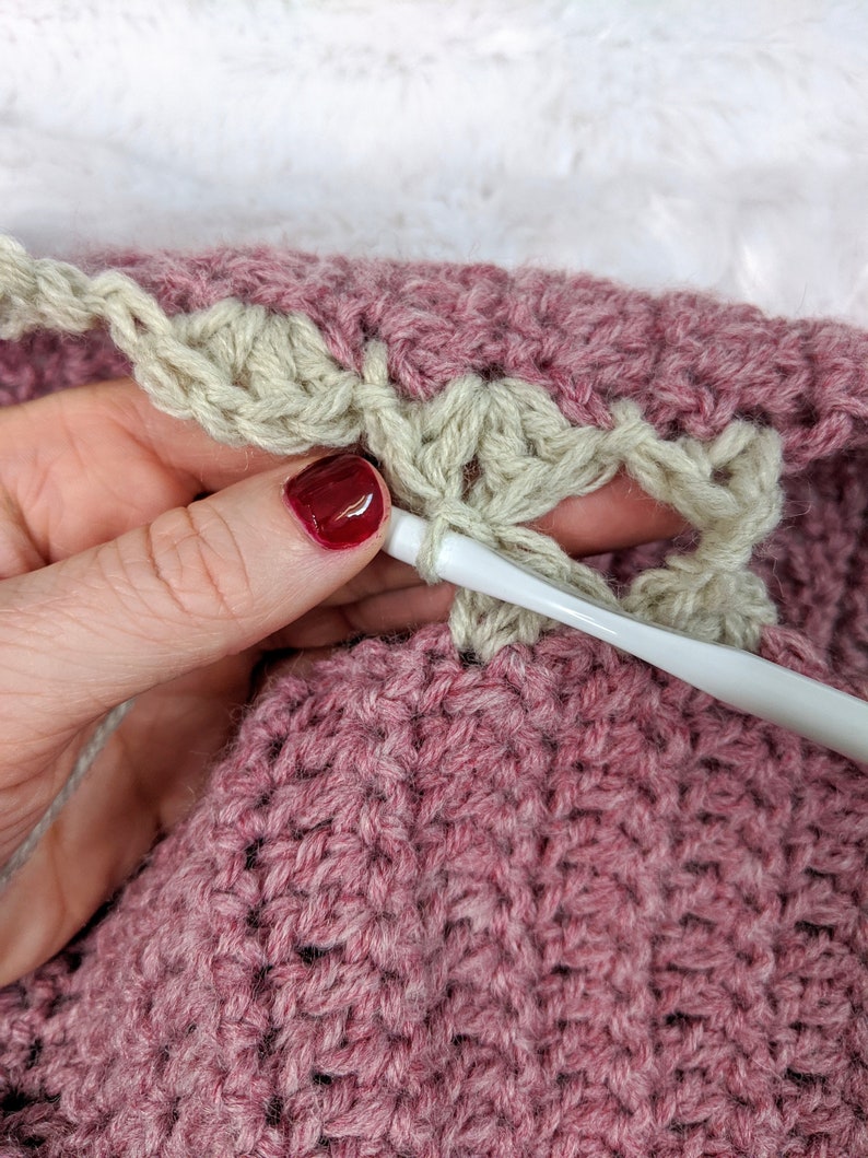 May include: Close-up of a person crocheting a pink and beige blanket. The person is holding a white crochet hook and is working on the beige border of the blanket.