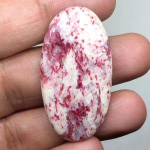 Peut inclure: Une pierre précieuse cabochon de forme ovale présentant une surface polie avec une base blanche et des inclusions rouge vif irrégulières dispersées sur toute la pierre. Le minéral affiche une texture cristalline avec des nuances de rouge.