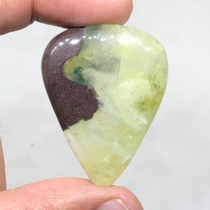 Puede incluir: Una piedra preciosa en forma de lágrima con una superficie pulida. La piedra presenta una mezcla de colores, incluyendo un marrón oscuro en la parte superior, que transiciona a un verde pálido y áreas translúcidas. La gema es sostenida por los dedos de una persona.
