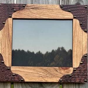 May include: A rectangular picture frame featuring a natural light brown wood border and dark brown faux alligator skin corners. The corners are secured with small, round, metallic studs. The frame contains a clear glass pane reflecting the outdoors.