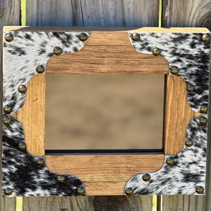 May include: Rectangular mirror with a wooden frame featuring cowhide accents and brass-colored nailhead trim. The mirror's central opening is surrounded by a light brown wooden frame with decorative cutouts. The cowhide has a black and white spotted pattern.