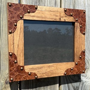 May include: A rustic wooden picture frame with a brown leather border and metal studs. The frame has a distressed wood finish and a rectangular shape.