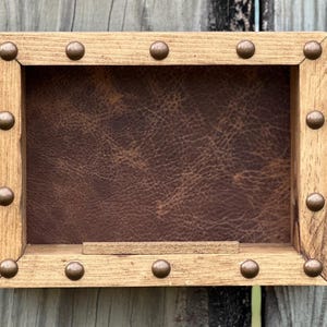 May include: A rectangular wooden shadow box with a brown leather backing. The frame is made of light-colored wood and is adorned with evenly spaced, round, bronze-colored studs. The shadow box is designed to display small items.