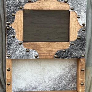 May include: A wooden picture frame with a mirror and a shelf. The frame is decorated with gray and white cowhide and brass studs.