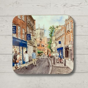 May include: A watercolour illustration of a street scene in a city. The street is lined with brick buildings, and there are people walking on the pavement. There is a church in the background, and the sky is blue. The illustration is printed on a coaster.
