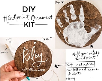 Könnte beinhalten: DIY-Handabdruck-Ornament-Set mit einem runden braunen Holz-Ornament mit dem Namen Riley und dem Datum Weihnachten 2022 in weißer Farbe. Das Set enthält einen vorgefertigten Namen und ein Datum, eine Schnur, Farbe und einen Pinsel.